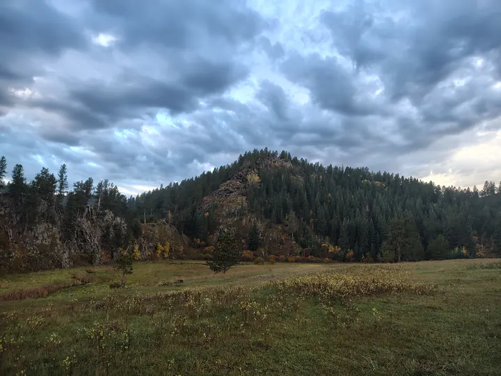 A grassy meadow beneath forested hills and a cloud-filled sky.